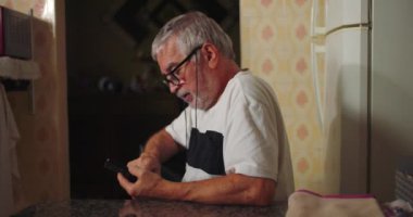 Yaşlı adam mutfak masasında oturuyor, telefonuna odaklanmış, gözlük takıyor, mutfak dekoruyla çevrili, sessiz teknoloji kullanıyor.