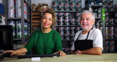 Gülümseyen adam ve kadın yerel küçük işletme nalburunda tezgahın önünde duruyorlar, etrafı güzelce düzenlenmiş boya ve alet raflarıyla çevrili, takım çalışması ve arkadaşlıkla dolu.