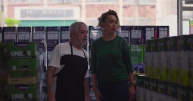 Kıvırcık saçlı, yaşlı bir erkek esnaf dükkanının koridorunda yaşlı bir erkek esnaf ile birlikte ürünleri inceleyen kadın müşteri, her ikisi de odaklı bir tartışma içindeler.