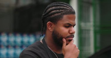 Düşüncelere dalmış genç siyahi bir adamın yan profili, çene üstüne el, yansıma, odaklanma ve profesyonel bir açık hava ortamında problem çözme zihniyeti.