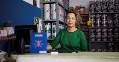 Hırdavatçıdaki bir masada oturan kadın girişimci, kendine güvenen ve yaklaşılabilir görünüyor, profesyonel bir perakende satış alanında boya ve malzeme raflarıyla çevrili.