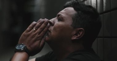 Duvara yaslanmış bir adam, dua jesti, başını yukarı kaldırmış, krizin ortasında inanç ve rehberlik arıyor, direnç, umut ve duygusal mücadele sunuyor.