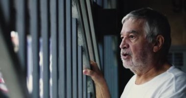 Metal parmaklıklar tutan yaşlı bir adam, garajdan caddeye bakıyor, düşünceli ve özenli bir şekilde kendini yansıtıyor.