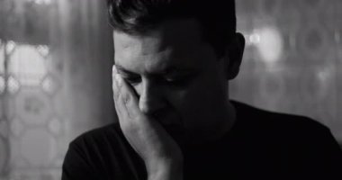 Siyah beyaz giyen bir adam, tek eli kapalı, gözleri kapalı, derin düşünce ya da üzüntü içinde, dantel perdeler ve sessiz bir atmosferde duvarlarla çevrili.