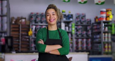 Tezgahın arkasında güvenle duran neşeli hırdavatçı, kolları çapraz, renkli malzemeler ve raflarda düzgünce yerleştirilmiş boya kutularıyla çevrili.