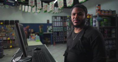 Boya dükkanında kasanın arkasında duran ciddi bir tezgahtar. Arka planda boya kutuları ve malzemeler görünüyor.