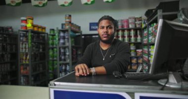 Siyah önlüklü bir adam boyacı tezgahına yaslanmış, raflar boya kutuları ve malzemelerle dolu, arka planda görünür, profesyonellik ve sakin tavır sergiliyor.