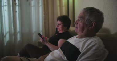 Son sınıf öğrencisi kadın telefonunda okuduğu içerikten dolayı kızgın. Kocasının yanında oturuyor ve televizyon izliyor. Duygusal zıtlığı vurguluyor.