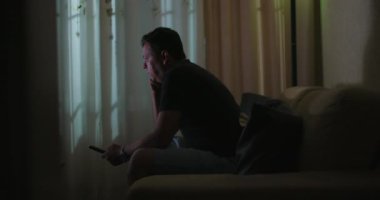 Televizyon seyrederken uzaktan kumandayı tutan, rahat bir şekilde koltukta oturan, arka planda perdeleri olan, ev eğlencesi, ev ortamında rahatlama ayarları yapan bir adamın samimi anı.