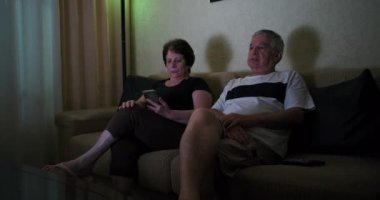 Kanepede oturan yaşlı çift, içeriği tartışırken televizyonu işaret eden adam, ikisi de endişeli ifadeler sergiliyor, akıllı telefon tutan kadın, kapalı alanda odaklanmış bir an.