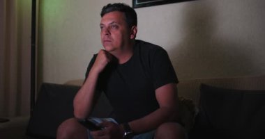 Kanepede oturup televizyon izleyen bir adam, elinde uzaktan kumandayla öne eğiliyor, odaklanmış ifade, evde rahat bir an, rahat bir oturma odası, ekranın içeriğiyle meşgul.