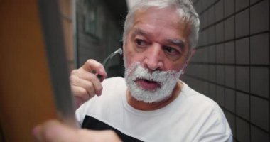 Evde köpükle tıraş olan yaşlı bir adam, düzgün bakım için jilet ve el aynası tutuyor, rahat bir ev ortamında kişisel bakım ve hijyene odaklanıyor.