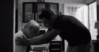 Oğlu, yaşlı babasını bunama ya da yaşlılıkla ilgili bir hastalıkla teselli etmek için mutfakta eğiliyor. Ellerini omzuna koyup şefkati ve duygusal bağlantıyı vurguluyor. Siyah ve beyaz.