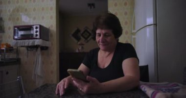 Son sınıf öğrencisi kadın mutfak masasında akıllı telefon kullanıyor. Ev ortamında günlük hayatın samimi ve rahat tasviri.