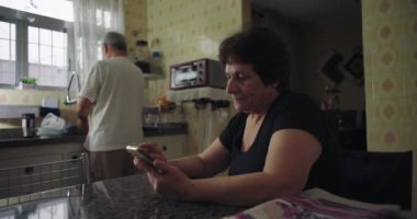 Mutfakta kıdemli bir çift, masada telefonu kontrol eden bir kadın kocası arka planda bulaşıkları yıkarken, rahat bir evde samimi bir şekilde günlük yaşantısını sürdürüyor.