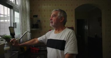 Mutfak penceresinin yanında duran yaşlı bir adam, düşünceli bir ifadeyle dışarı bakıyor, doğal bir iç mekan düzeni, ev hayatındaki sıradan bir an, samimi ve düşünceli.