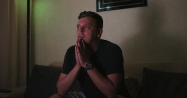 Şok edici TV içeriğine tepki veren, elleriyle yüzünü sıkan, şaşı gözlü, yoğun bir ilişki, duygusal gerilim, kapalı alanda dramatik bir an yaşayan adam.