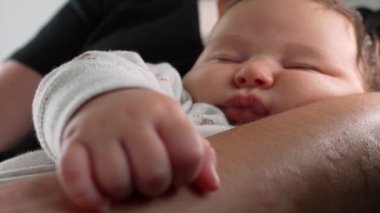 Bebeğin küçük eli ebeveynin kolunda yavaşça dinleniyor, bebek huzur içinde uyuyor, bir aile ortamında sıcaklık, sevgi ve güven hissi yaratıyor. 