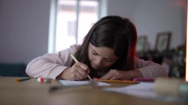 Çocuk, oturma odasındaki masada, sanatsal bir meşgaleyle uğraşırken konsantre bir ifadeyi gösteren kağıda gömülür..