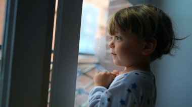 Sabahları pijama giyen küçük bir çocuk pencereden dışarı bakıyor.