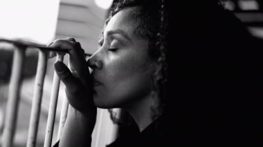 Üzgün, kederli, siyah bir kadın depresyondan gözleri kapanıyor balkondan metal çubuğa yaslanarak evde dramatik siyah beyaz monokromla sessiz bir umutsuzlukla mücadele ediyor.