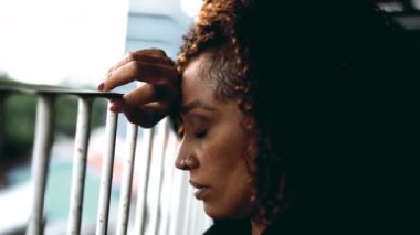 Hüzün ve umutsuzluk hisseden depresif siyah bir kadın. Gözleri kapalı. Balkonda metal bir bara yaslanmış.