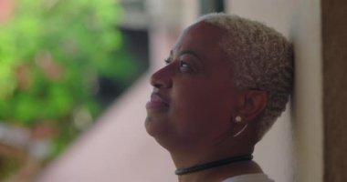 Latin kökenli, kısa kıvırcık saçlı, açık havada duvara yaslanmış, gözleri açık, sakin ve içe dönük, huzurlu bir anda yukarıya bakan Latin bir kadın.