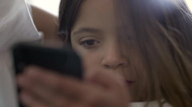 Küçük bir kız cep telefonu aygıtı kullanıyor. Ekrana bakıyor. Modern teknolojiyle meşgul olan çocuğun sıkı yüzü çevrimiçi medyaya bakıyor.