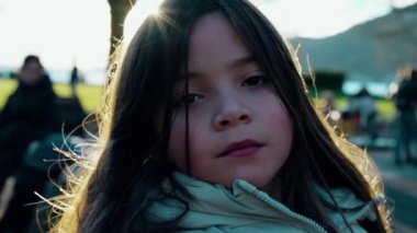 Gün batımında oyun parkında, soğuk mevsimde ceketini giymiş, arka planda bulanık insanlar olan bir çocuğun yakın çekim yüzünü izleyen vakur küçük kız.