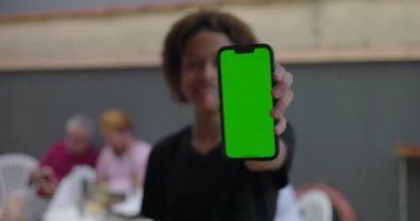Genç, yeşil ekranlı akıllı telefonu tutuyor, kendine güvenerek gülümsüyor, çeşitli yetişkinlerin bulanık arka planda etkileşim kurduğu sıradan bir toplantıda.