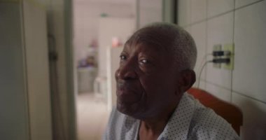 Afrika kökenli yaşlı bir adam fayanslı mutfakta oturuyor, kameraya nazikçe gülümsüyor, sıcaklığını ve bilgeliğini iletiyor, ev ortamında her gün samimi anlar yaşıyor.