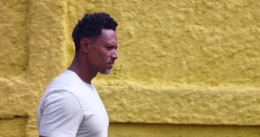 Sarı duvar boyunca yürüyen bir adam sokak kenarı profilinde düşünceli bir ifadeyle Afrika kökenli bireysel yansıtıcı ve düşünceli bir yaşam tarzıyla
