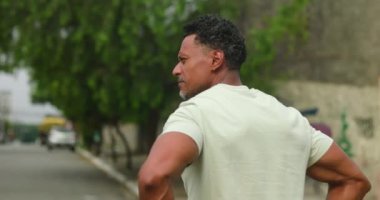 Bir adam durup, düşünceli bir ifadeyle şehir caddesinde dönüp duruyor. Çevreyi inceliyor yeşillik ve binalar arka planda Afrika kökenli bireyler.