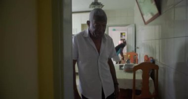 Yaşlı bir siyah adam mutfaktan oturma odasına yürüyor, tarafsız bir ifade takınıyor, ev içinde görünür mobilyalar ve dekorla geçiş yapıyor.