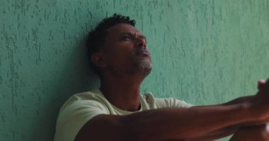 Afrika kökenli bir adamın yeşil bir duvara yaslanmış, düşünceli bir ifadeyle yukarıya bakan sessiz bir yalnızlık, yansıtıcı bir an, zihinsel sağlık farkındalığı.