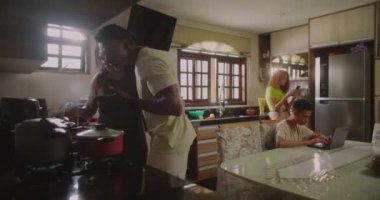 Baba mutfağa girer, karısını sevgiyle öper, genç kız yemek masasında dizüstü bilgisayarda çalışırken onu öper ve anne yemek pişirmeye devam eder.