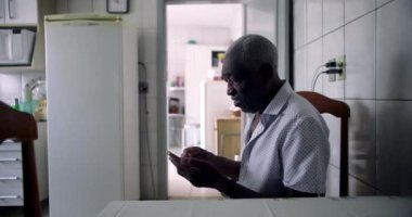 Rahat bir mutfakta yemek masasında oturan yaşlı Afrikalı Amerikalı adam akıllı telefon kullanmaya odaklanmış ve günlük kullanım anlarını gösteriyordu.