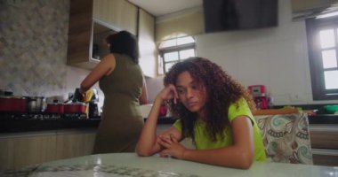 Anne tezgahta yemek pişirirken kıvırcık saçlı genç kız yemek masasına yaslanıyor, hayal kırıklığına uğramış görünüyor, aile dinamikleri evde.