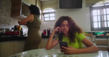 Genç kız yemek masasında telefona odaklanmış, anne arka planda mutfakta yemek pişiriyor, günlük aile rutini, Afrika kökenli aileler...