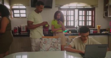 Mutfakta aile bağları, anne yemekleri, telefonda kızının yanında duran baba, yemek masasında dizüstü bilgisayara odaklanmış genç bir çocuk, günlük yaşam ve bağlantı, Afrika kökenli bir aile.