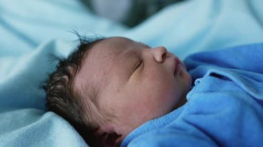Mavi kıyafetli yeni doğmuş bir bebeğin yan profili, mışıl mışıl uyuyor. Bebeğin huzurlu ifadesi, güzel saçları ve yumuşak teni, yeni doğmuş bir bebeğin masumiyetini ön plana çıkarıyor.