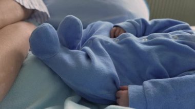 Mavi tulumun içinde dinlenen yeni doğmuş bir bebeğin yakın çekimi, tatlı ve huzurlu bir uyku anı. Bebek yumuşak mavi bir battaniyeye uzanıyor, erken yaşlardaki sükuneti ve saflığı vurguluyor.