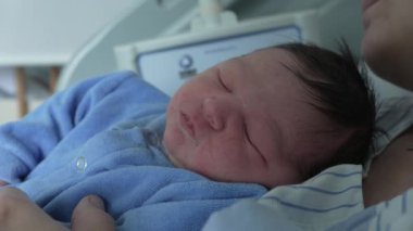 Mavi tulumlu yeni doğmuş bir bebeğin yakın plan çekimi, anne babanın göğsünde huzurlu bir şekilde uyuması, bir hastane ortamındaki derin bağı ve huzurlu çocukluk anılarını vurguluyor.