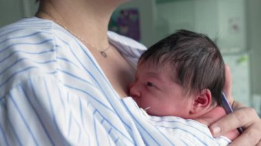Anne hastane ortamında bebeğini göğsüne yakın bir şekilde kucaklıyor. Bebek çizgili bir tişörte sarılmış, ten tene teması vurguluyor, anne ve çocuk arasındaki hassas bağı gösteriyor