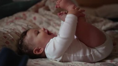 Bebek yatakta uzanıyor, gülümsüyor ve ayakları tutuyor, sıcak ve samimi bir ev ortamında eğlenceli ve ilginç hareketler yapıyor.