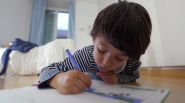 Yerde yatan genç bir çocuk, mavi bir kalemle çiziyor, odaklanmış bir ifade, derin bir konsantrasyon içinde, yaratıcı öğrenme ve sanatsal ifade ile bir ev ortamında meşgul