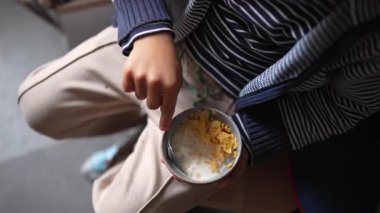 Çocuk elinde sütle bir kase mısır gevreği, kaşıkla kaseye batırılmış, yukarıdan yakalanan sıradan kahvaltı anı, basitliği ve besinleri vurguluyor.