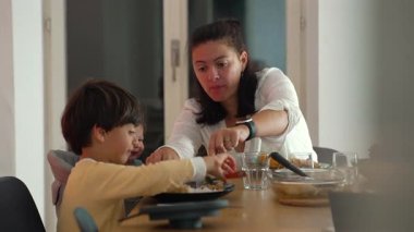 Anne, yemek masasında küçük çocuklara yemek kesiyor, aile yemekleri sırasında öğretiyor ve yetiştiriyor, evde samimi bakım ve etkileşim anları yaşıyor.