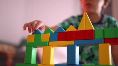 Genç çocuğun elinin, renkli ahşap yapıya mavi üçgen blok yerleştirmesi, oyun zamanı odak ve yaratıcılığı vurgulaması, kapalı alanlarda yaratıcı aktiviteleri keşfetmesi,