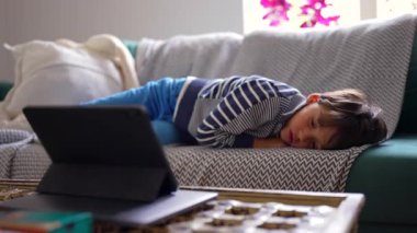 Genç bir çocuk kanepede uzanmış, tablet ekranı önünde, dinleniyor ve izliyor, rahat bir ev ortamında, rahat ve rahat bir çocuk, düşünceli bir ifade, huzurlu bir ortam.